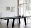 Minimalist modern dining room with a sleek black wooden dining table featuring angular legs, complemented by white chairs, soft natural lighting from large windows, and simple wall art in a serene white interior.