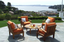 Outdoor patio set with four wooden chairs featuring vibrant orange cushions, arranged around a round wooden table with drinks and snacks, overlooking a scenic bay and marina.