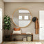 A stylish entryway featuring a large round wooden-framed mirror above a rustic bench with decorative pillows. A coat rack with a hat and jacket, a potted plant, and woven baskets add warmth and functionality.